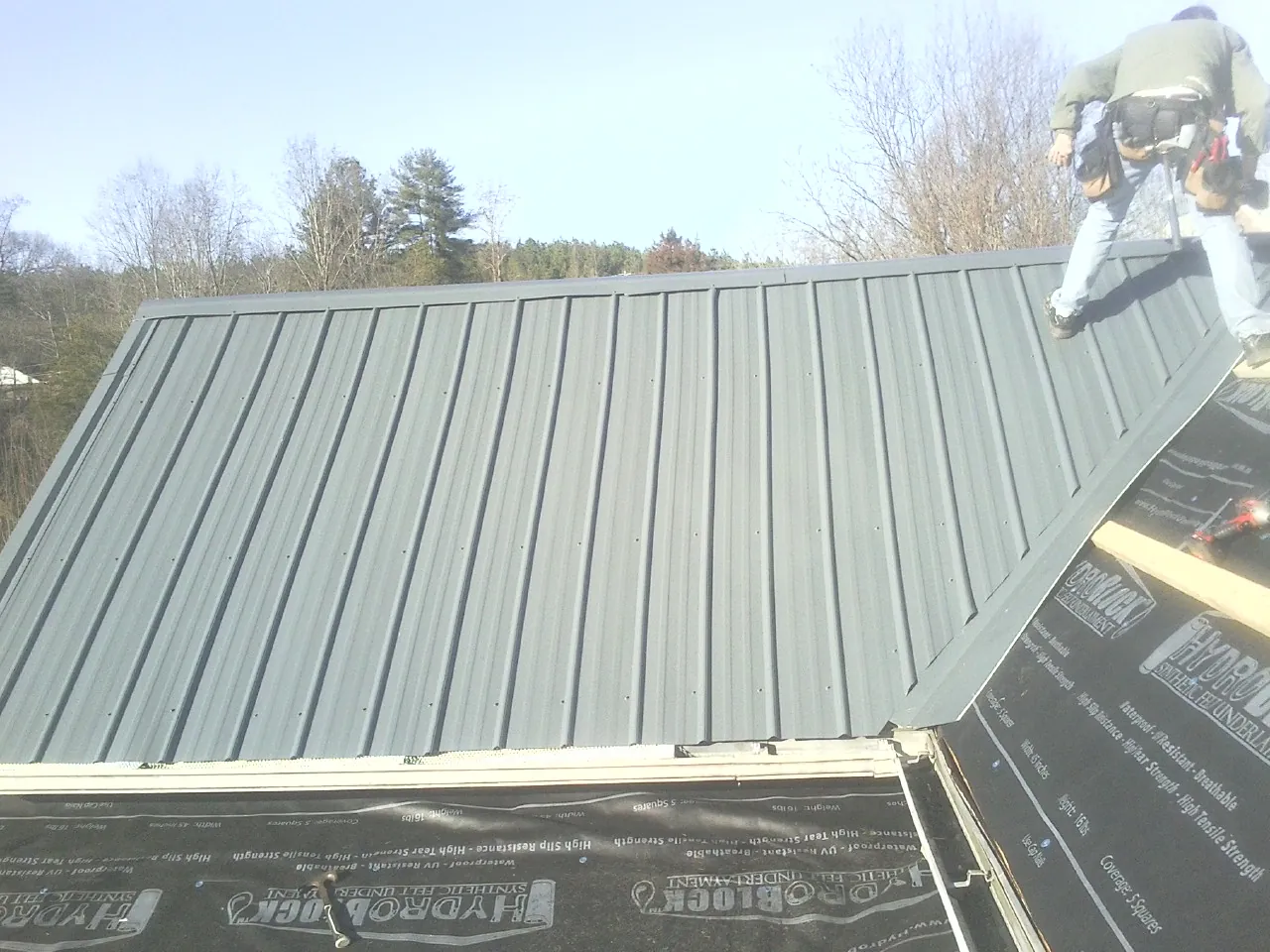 Crew installing dark green standing seam panels over synthetic underlayment in Rego Park