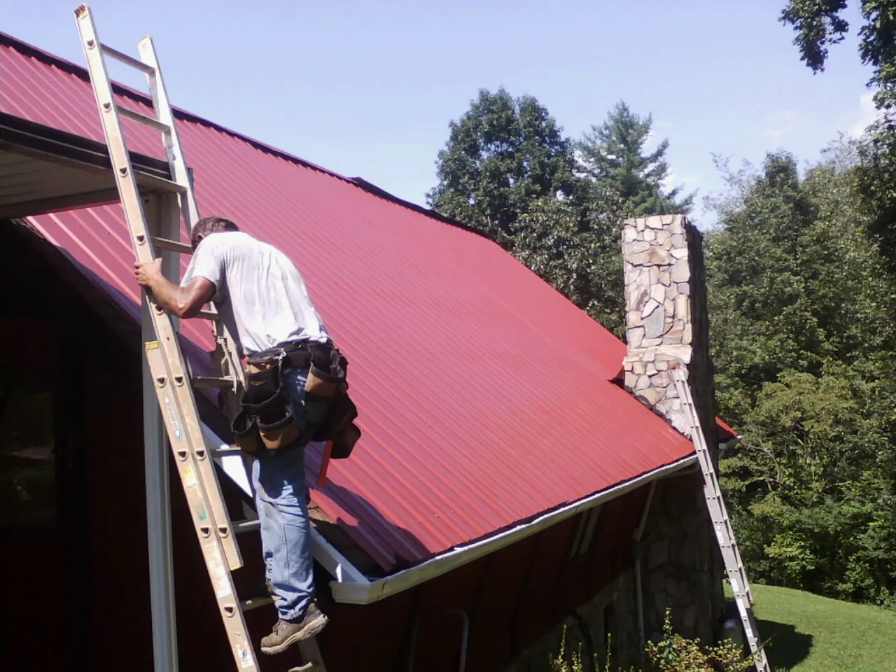 Roofer on a ladder beside a red metal roof and stone chimney — Soffit & Fascia Repair by Roofing Brothers in Rego Park